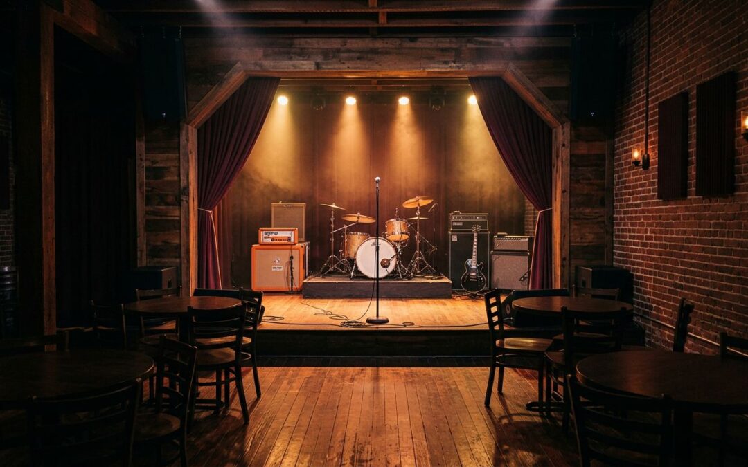 A brick music venue with an empty stage, instruments, tables, and warm amber lighting.